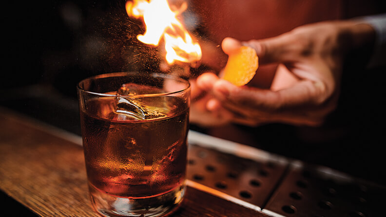 A glass of liquor on a bar with a bartender lighting a flame behind it.